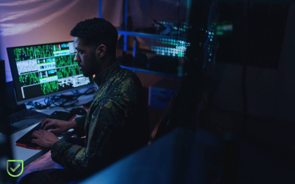 Soldado en uniforme de camuflaje trabajando en una laptop con códigos en el fondo, representando la guerra cibernética