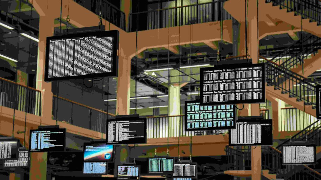 Varios monitores suspendidos en un entorno industrial muestran líneas de código en texto blanco sobre fondo negro. La escena resalta la importancia del monitoreo y la seguridad en tiempo real en sistemas que utilizan Linux, planteando la pregunta de si se necesita antivirus para Linux en estos entornos.